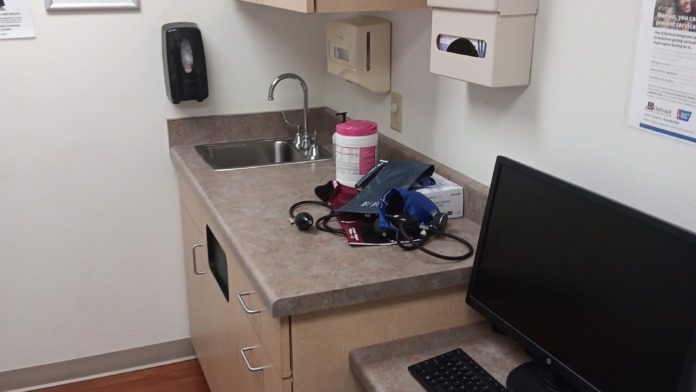 Interior clinical workstation with countertop sink, computer setup, medical equipment storage, and health education materials on wall