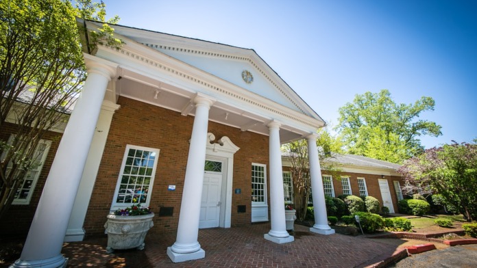 Exterior front entrance of brick building with white columns, pediment roof, manicured landscaping, and brick walkway