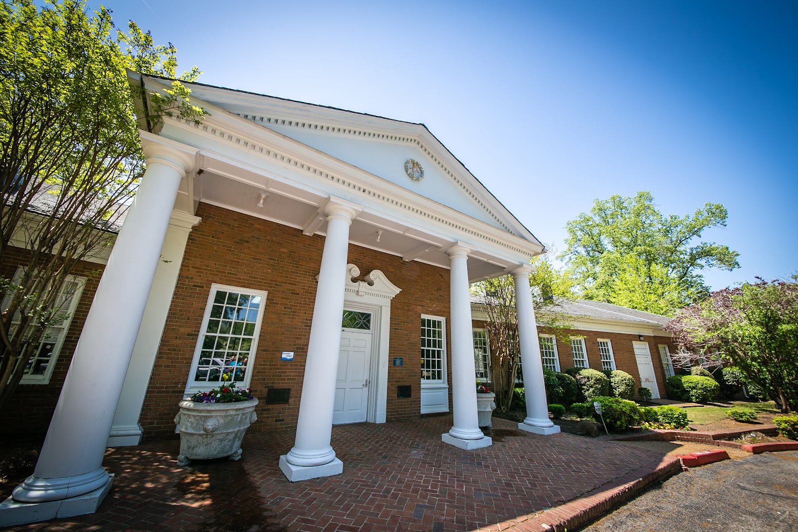 Exterior front entrance of brick building with white columns, pediment roof, manicured landscaping, and brick walkway