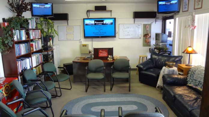 Interior common room with seating areas, desks, bookshelves with plants, television monitors, and ambient lighting