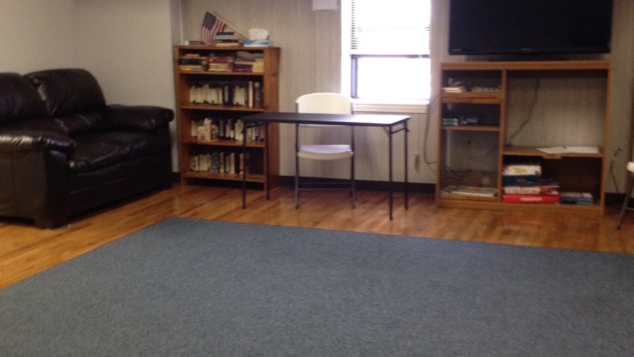 Interior common room with leather sofa, wooden shelving, mounted television, desk, and hardwood flooring.