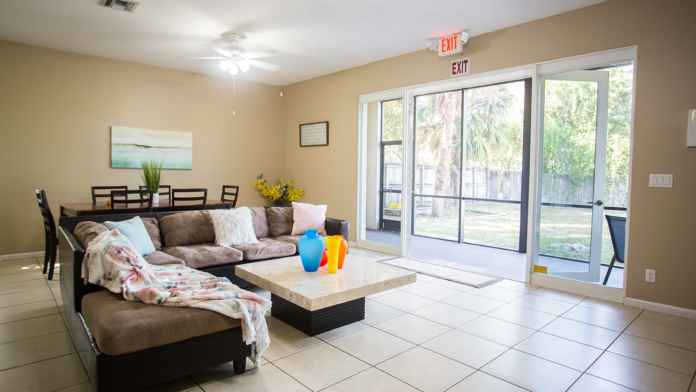Interior community space with beige walls, brown sofa with pillows, dining furniture, TV, and large glass doors opening outside.