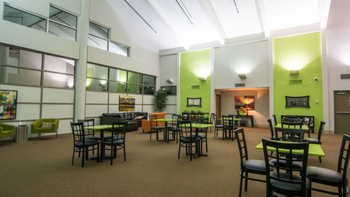 Interior communal space with lime green and white walls, multiple dining tables with black chairs, sofas, and natural ceiling skylights