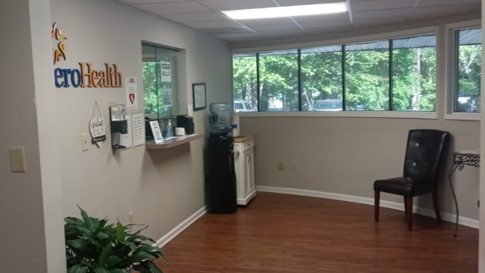 Interior common area with SperoHealth logo, extensive windows overlooking trees, black seating chair, desk area, and plant decoration