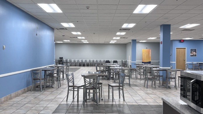 Interior dining room with multiple round and rectangular tables, chairs, blue accent walls, serving area, and natural lighting from skylight panels