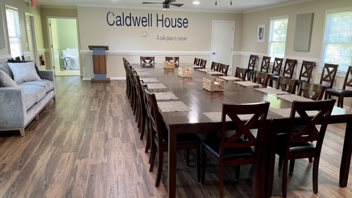 Interior dining room with multiple wooden tables, chairs, and floor-to-ceiling windows providing natural light, featuring Caldwell House signage on wall