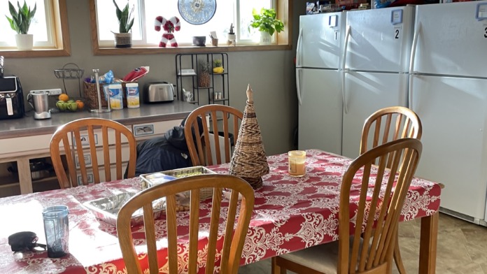 Interior dining area with wooden dining table, multiple chairs, kitchen appliances, and large windows with natural light