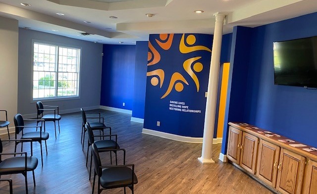Interior group therapy room with rows of chairs, blue accent walls with Spero Health logo, wooden media console, and natural window lighting