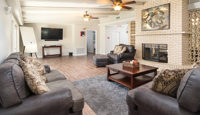 Interior community living room with gray seating, fireplace, television, and natural lighting from windows