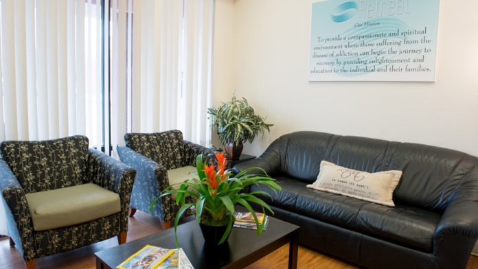Interior common lounge space featuring patterned upholstered armchairs, black leather sofa, decorative plants, and Retreat mission statement wall display