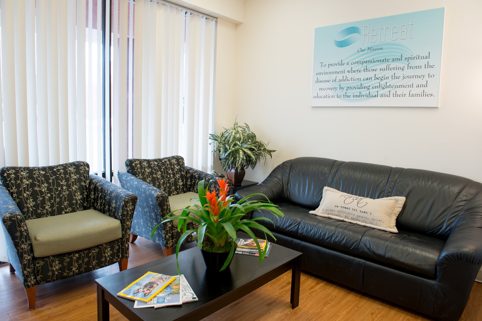 Interior common lounge space featuring patterned upholstered armchairs, black leather sofa, decorative plants, and Retreat mission statement wall display