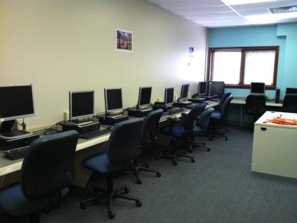 Interior computer room with multiple desktop workstations, chairs, and desks along walls