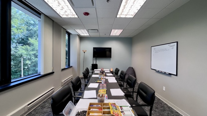 Interior conference room with long table, black chairs, presentation screen, whiteboard, and large windows
