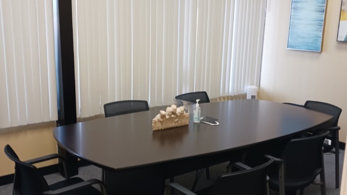 Interior meeting room with large oval table, black chairs, windows with vertical blinds, and wall art