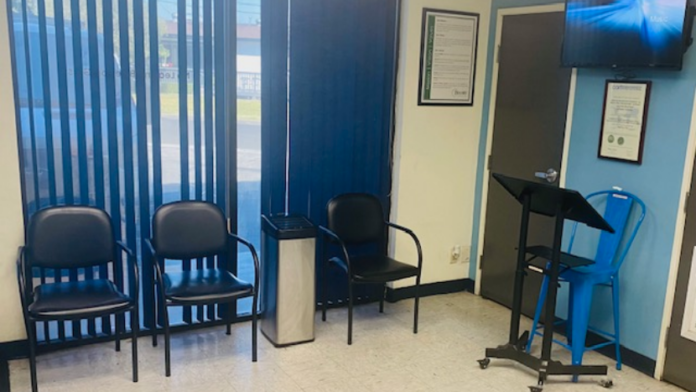 Interior consultation room with vertical blinds on windows, blue and black chairs, wall-mounted television, and framed documents.