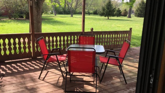 Exterior covered deck with red chairs, round table, decorative railing, and expansive green grounds in background