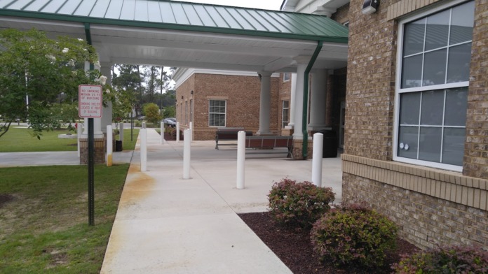 Exterior covered entrance with white support columns, green metal roof, brick building, and landscaped grounds