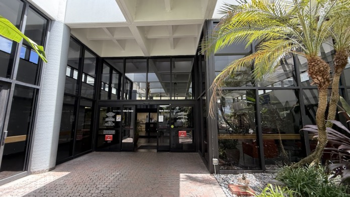 Exterior covered portico entrance with architectural white beams, tropical plants and palm trees, paved walkway, and facility entry