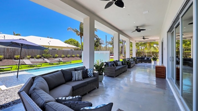 Interior patio space with black furniture, ceiling fans, glass doors, pool view, and landscaped surroundings visible through openings.