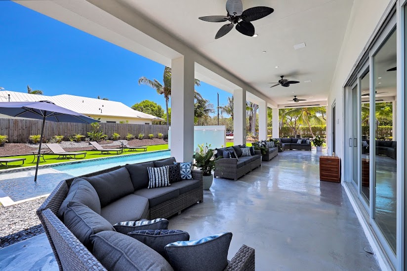 Interior patio space with black furniture, ceiling fans, glass doors, pool view, and landscaped surroundings visible through openings.