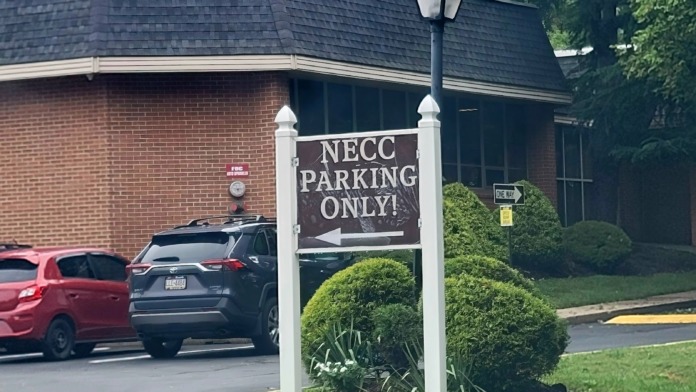 Exterior parking lot area with NECC parking only sign, brick building wing, landscaping, and vehicle parking spaces