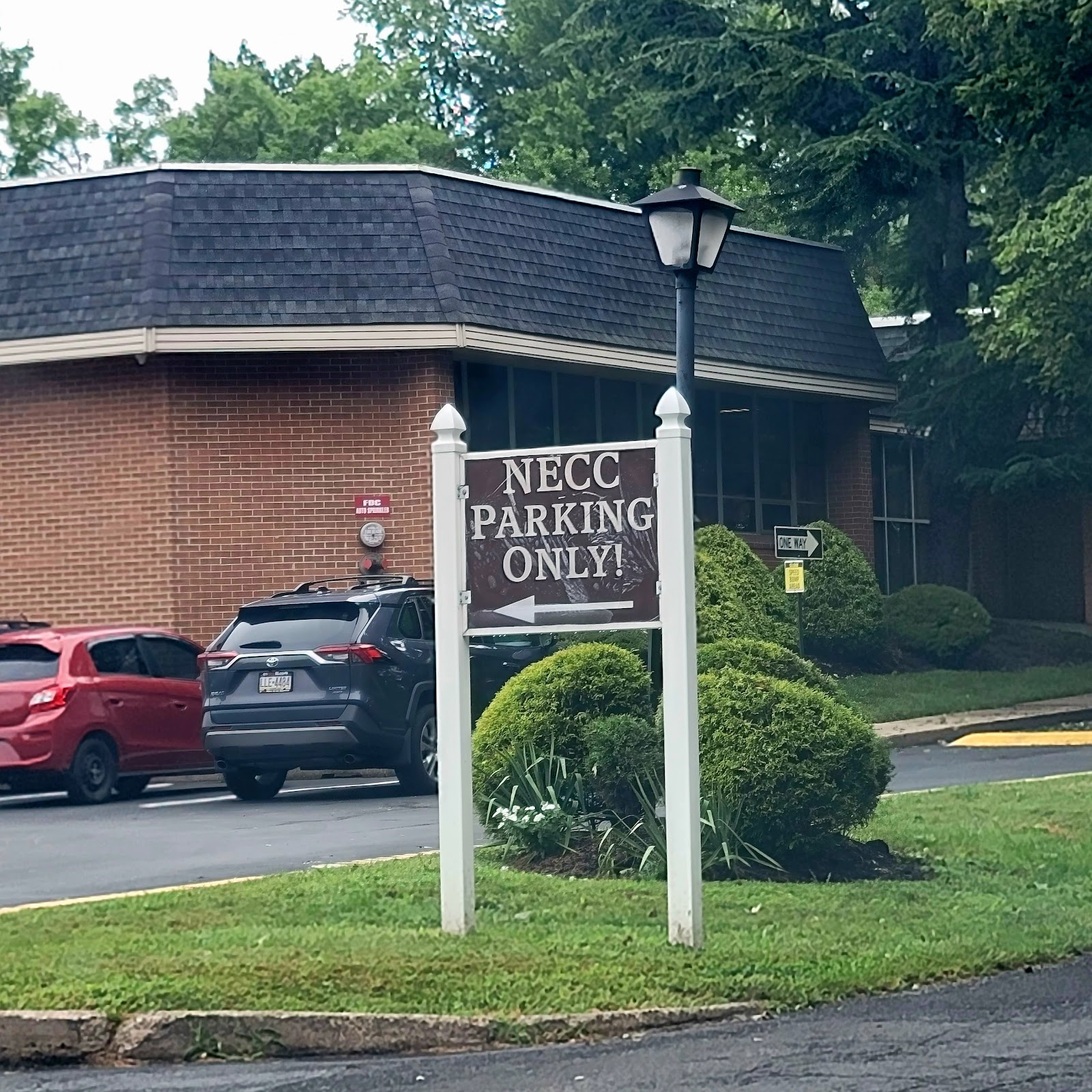 Exterior parking lot area with NECC parking only sign, brick building wing, landscaping, and vehicle parking spaces