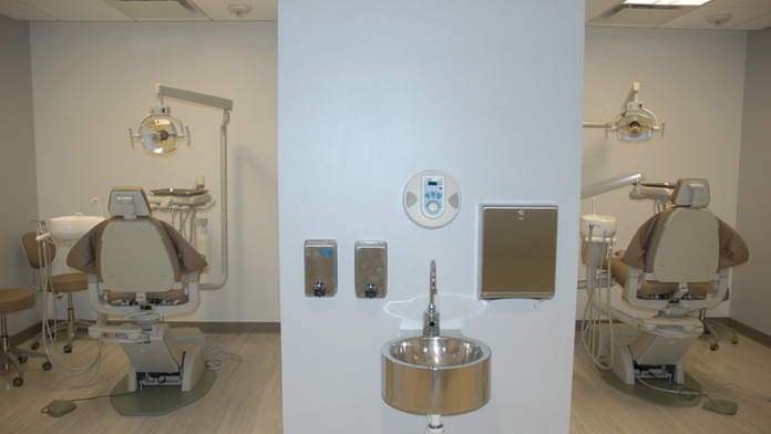 Interior dental treatment facility with two professional chairs, overhead lights, sink, and wall-mounted equipment