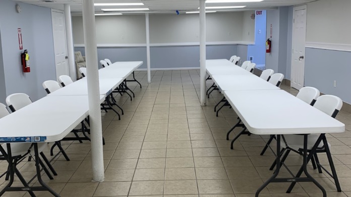 Interior dining hall with long white tables, black folding chairs, tile flooring, and fluorescent ceiling lighting