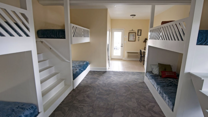 Interior dormitory room with multiple built-in bunk beds, blue bedding, carpeted floor, and connecting bathroom area