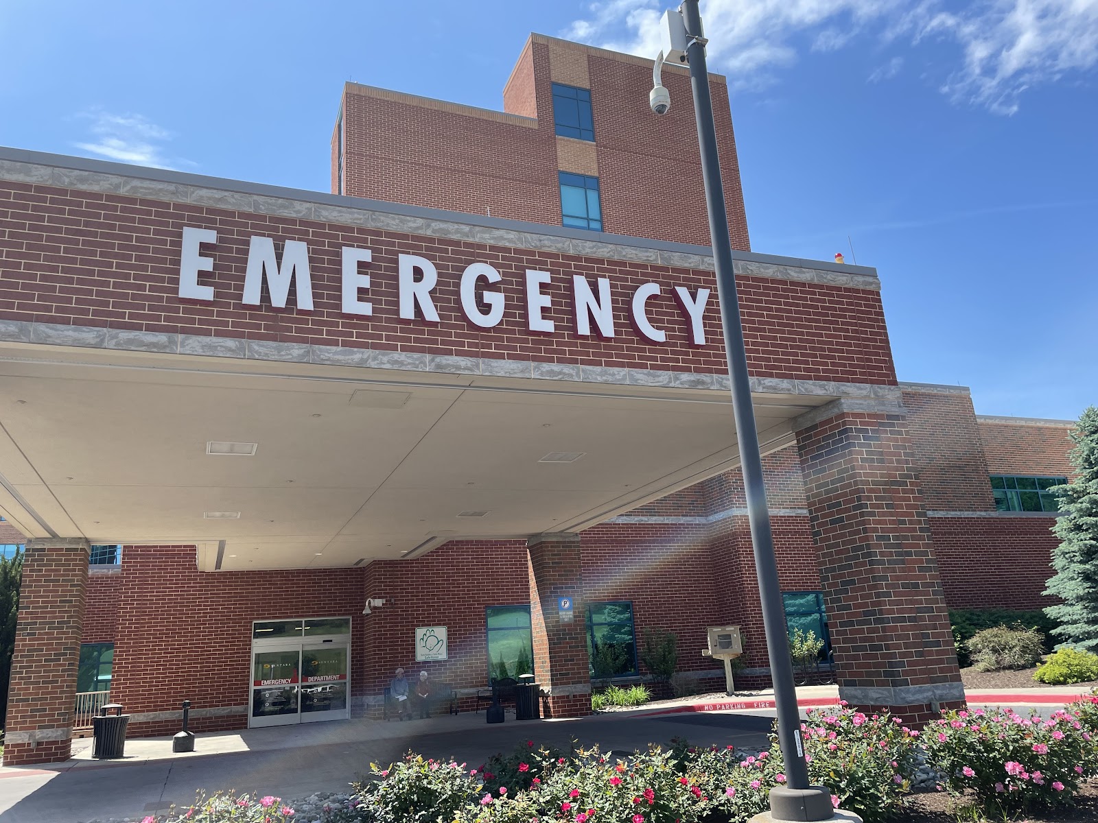 Emergency department entrance with signage, brick exterior, covered portico, and landscaped plantings with flowering shrubs