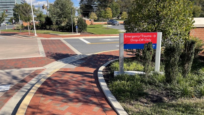 Exterior view of emergency drop-off area with directional signage, paved driveway, landscaped grounds, and parking area