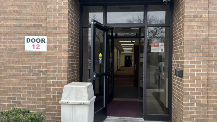Exterior entrance with black frame glass door labeled Door 12, concrete walkway, and brick building facade