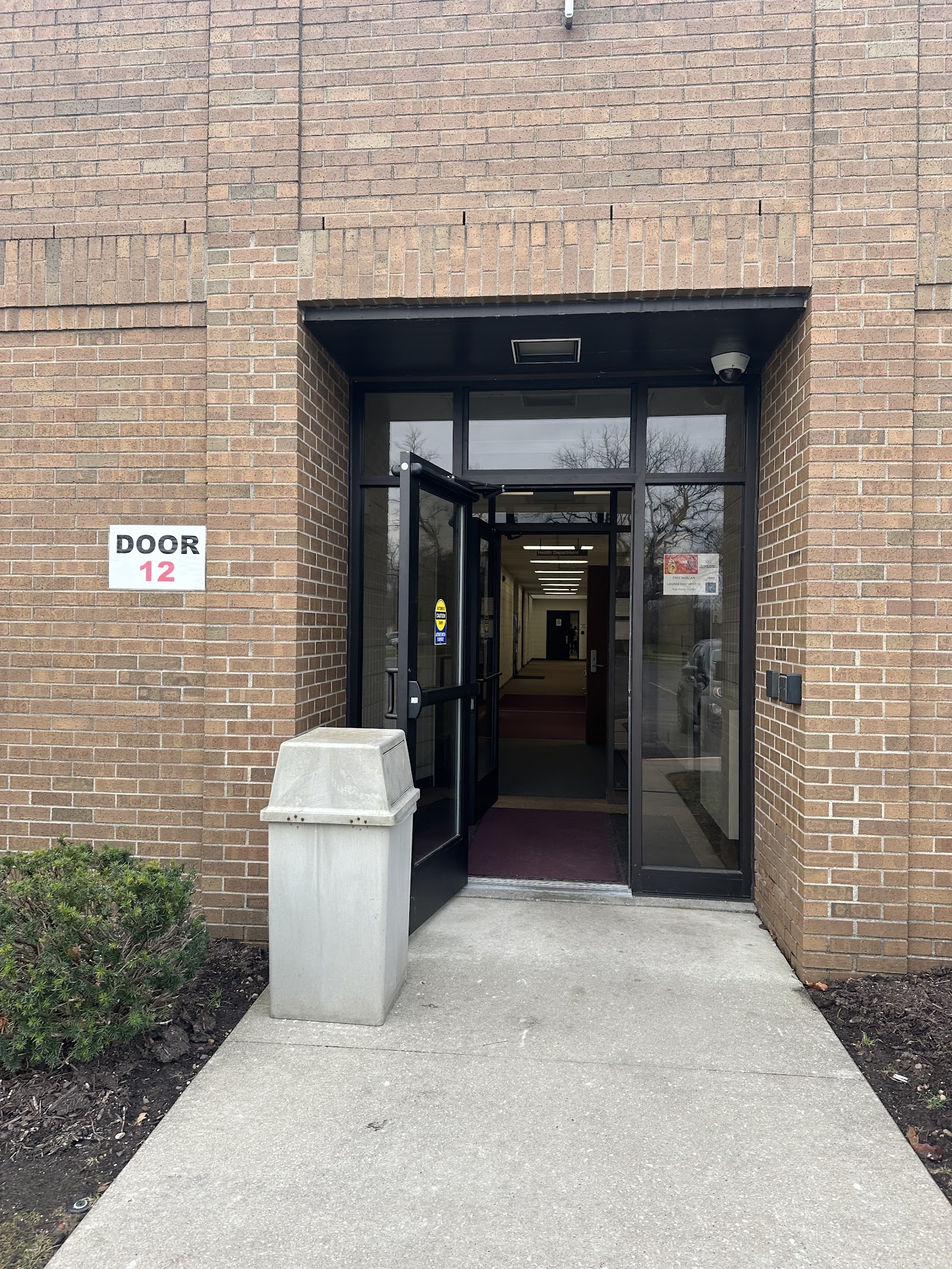 Exterior entrance with black frame glass door labeled Door 12, concrete walkway, and brick building facade