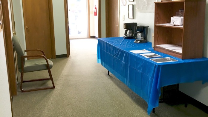 Interior multipurpose room with blue-draped table, wooden furniture, shelving unit, exit signage, and doors to adjacent spaces
