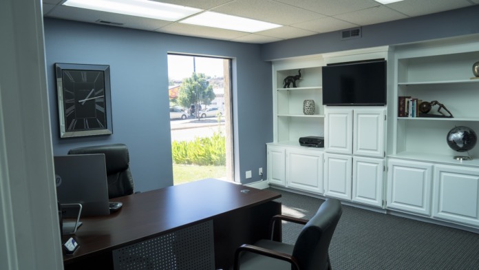Interior executive office with dark desk, computer equipment, wall clock, shelving units, and large windows