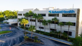 Exterior multi-story white facility building with AdventHealth signage, palm trees, and parking area from elevated street view