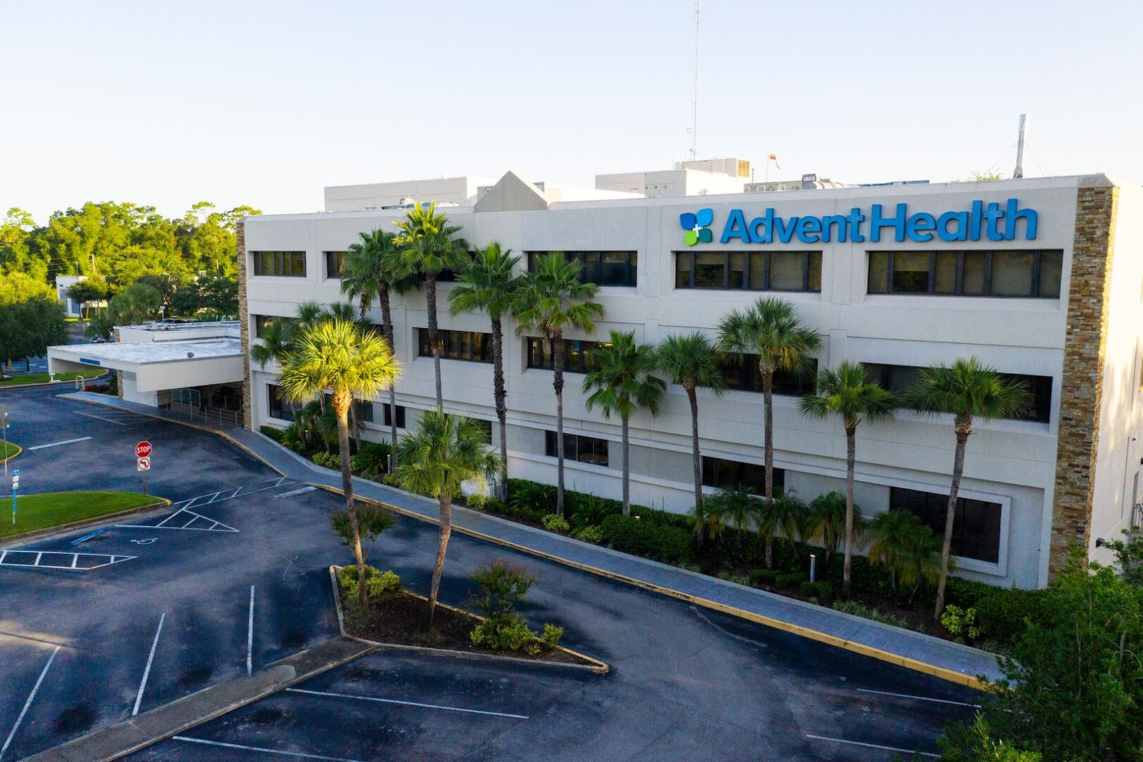 Exterior multi-story white facility building with AdventHealth signage, palm trees, and parking area from elevated street view