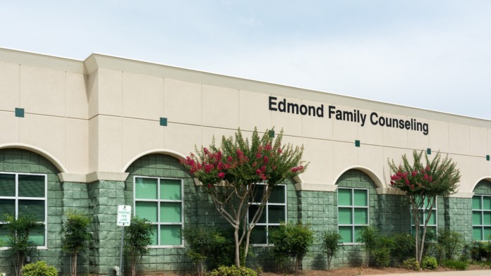 Exterior building view showing main entrance doors, landscaping with trees and shrubs, and parking area approach