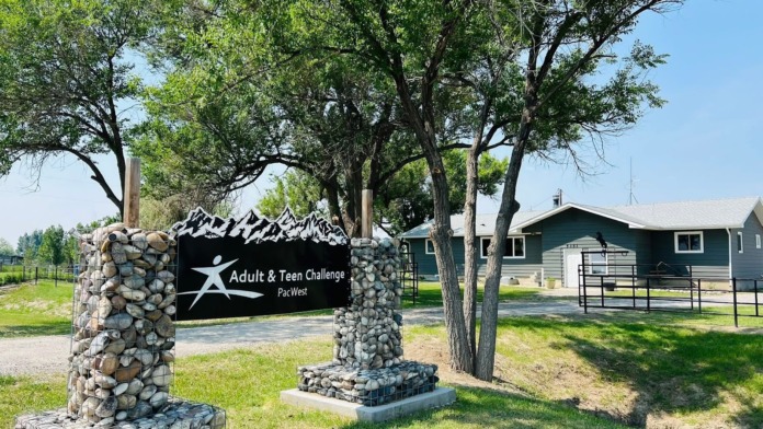 Exterior view of facility grounds showing stone entry sign with branding, green landscaping, and residential buildings in background