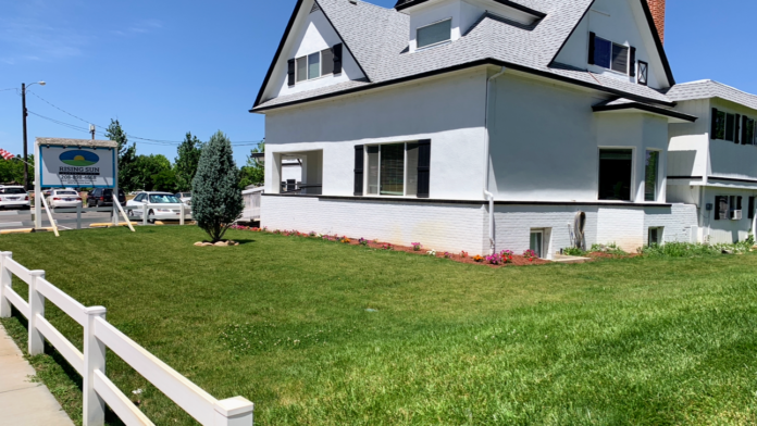 Exterior view of white multi-level facility building with manicured lawn, flower beds, and white fencing along street