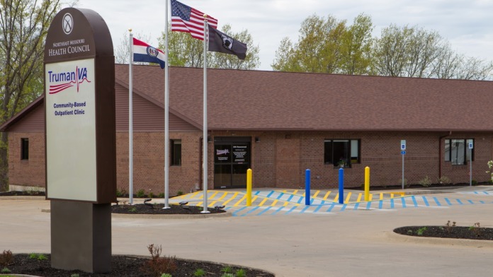 Exterior view of brick medical facility showing main entrance, facility signage, flagpoles, and accessible parking.