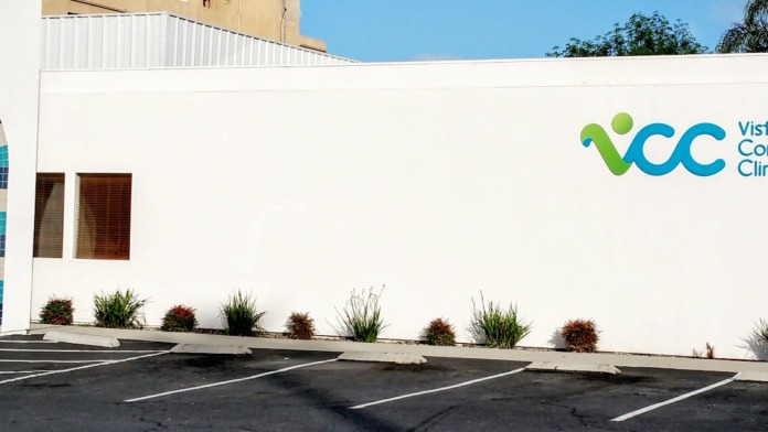 Exterior view of white clinical building showing decorative tiled archway entrance, landscaping, and parking area in sunny conditions