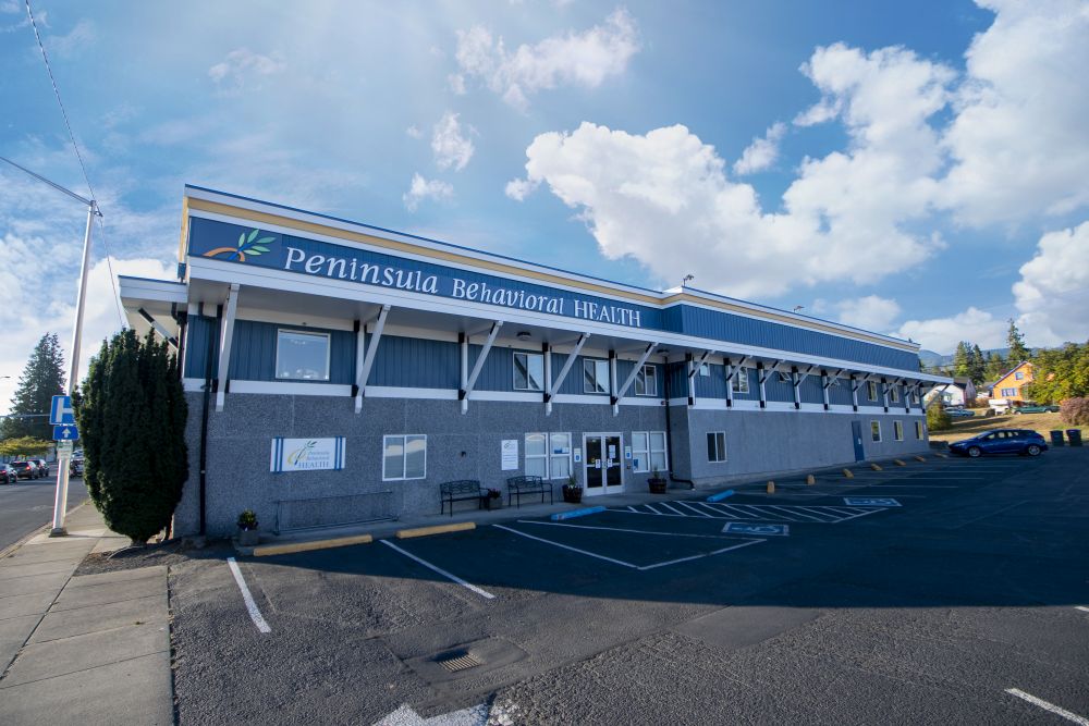 Exterior view of Peninsula Behavioral Health building showing blue roof, main signage, parking spaces with handicap access, and surrounding neighborhood