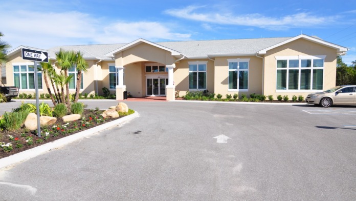 Exterior view of single-story beige building with large windows, palm landscaping, and parking area with one-way sign