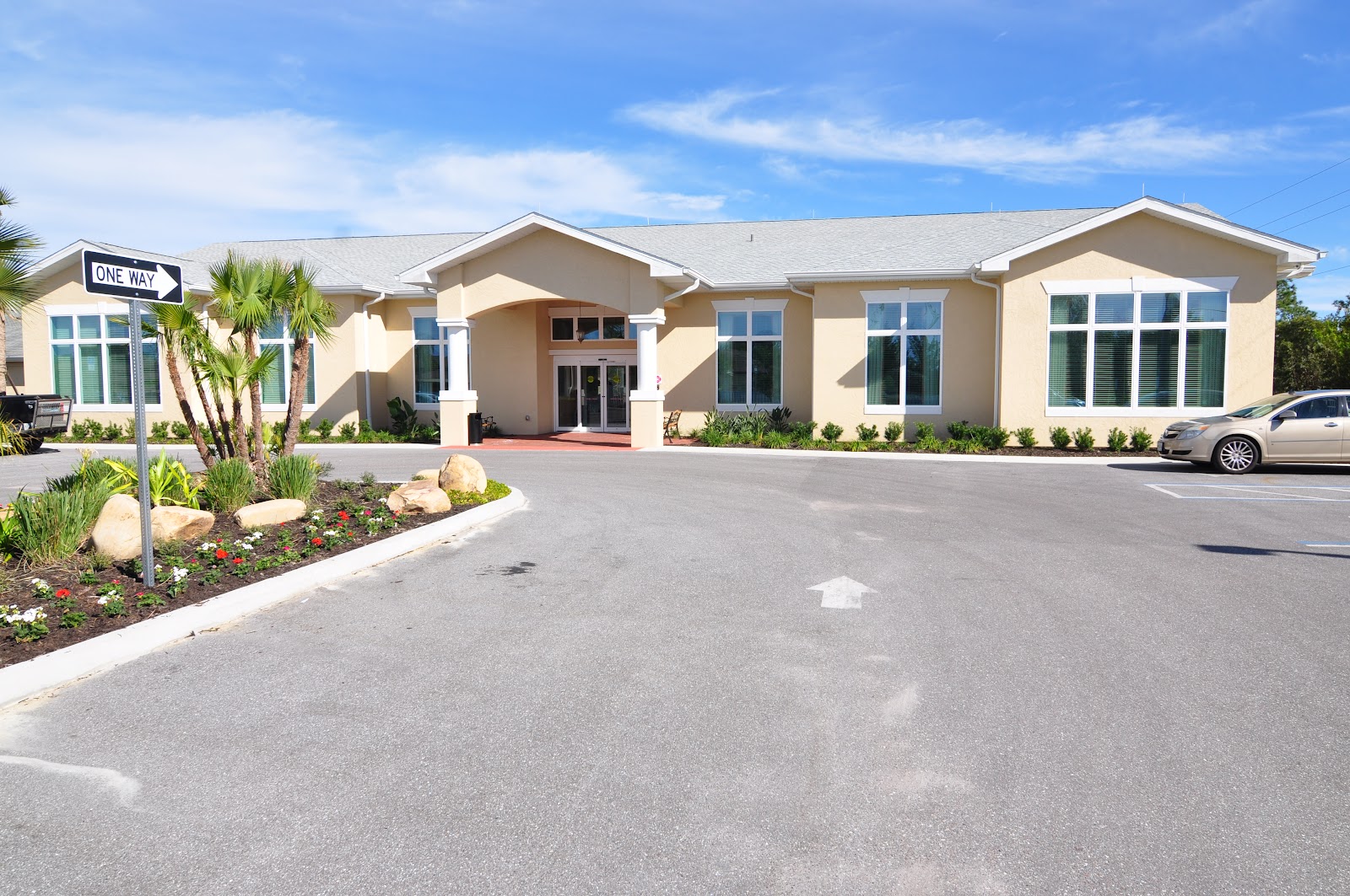 Exterior view of single-story beige building with large windows, palm landscaping, and parking area with one-way sign