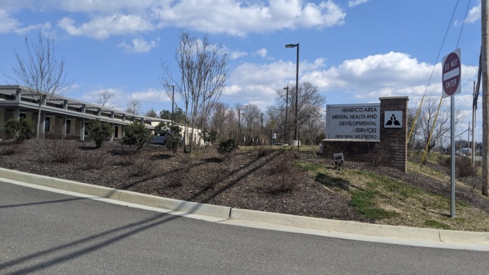 Exterior approach to facility showing brick building, landscaped grounds with trees, directional signage, and parking access
