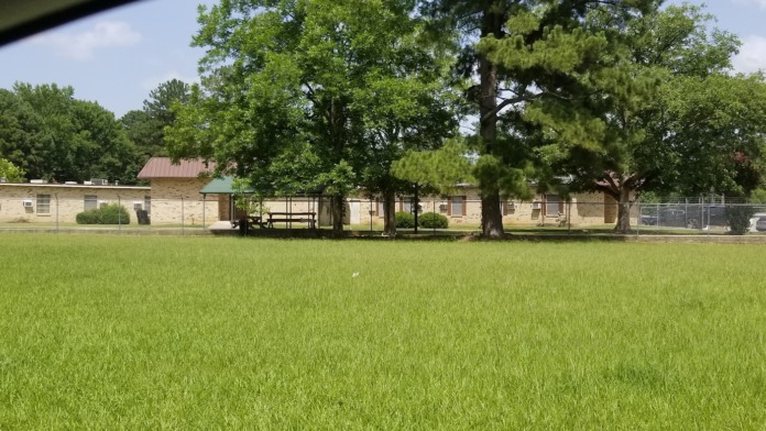 Exterior view of stone facility building across manicured lawn with mature shade trees and fenced perimeter