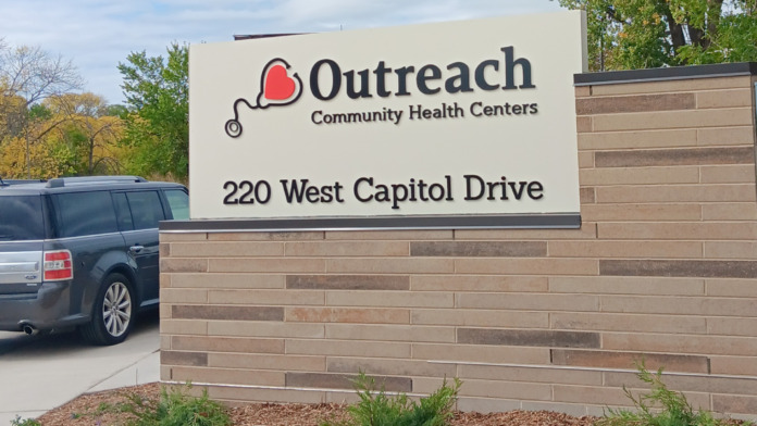 Exterior sign displaying Outreach Community Health Centers name and address at 220 West Capitol Drive with parking area and landscaping