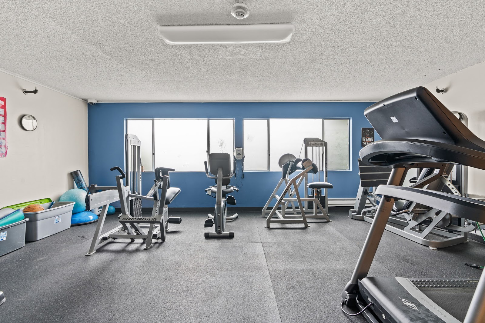 Interior fitness center with exercise equipment including treadmills, stationary bikes, blue accent wall, and large windows