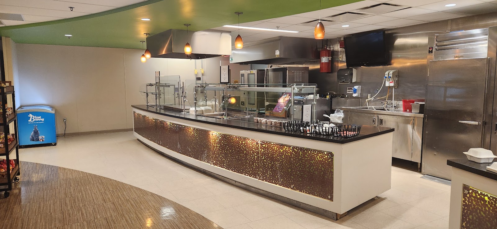 Interior commercial kitchen with stainless steel equipment, serving counters, pendant lighting, and modern food preparation area